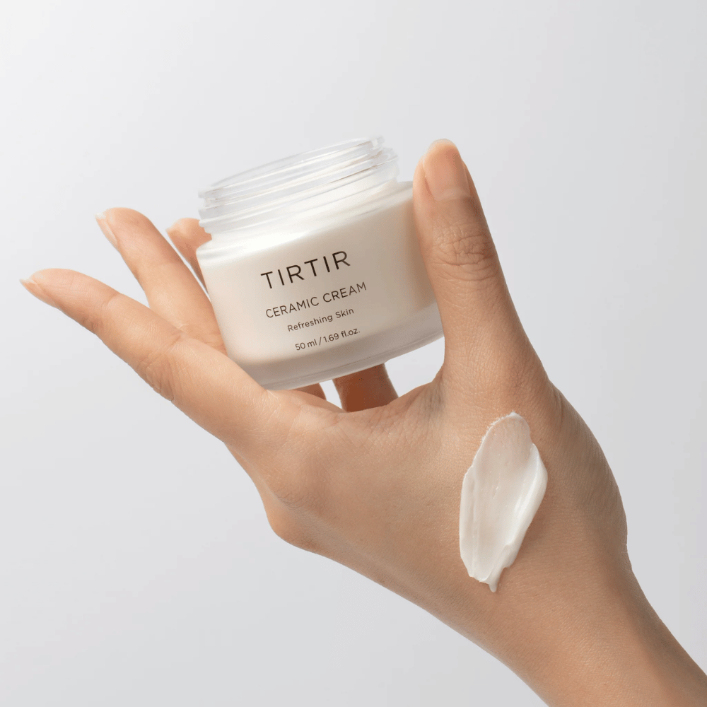 Uma mão segura um frasco com TIRTIR Ceramic Cream. No dorso da mão, uma quantidade do creme foi aplicada. O creme parece ser espesso e hidratante. A embalagem indica que o conteúdo é para "Refreshing Skin" e a quantidade é de 50 ml / 1,69 fl oz.