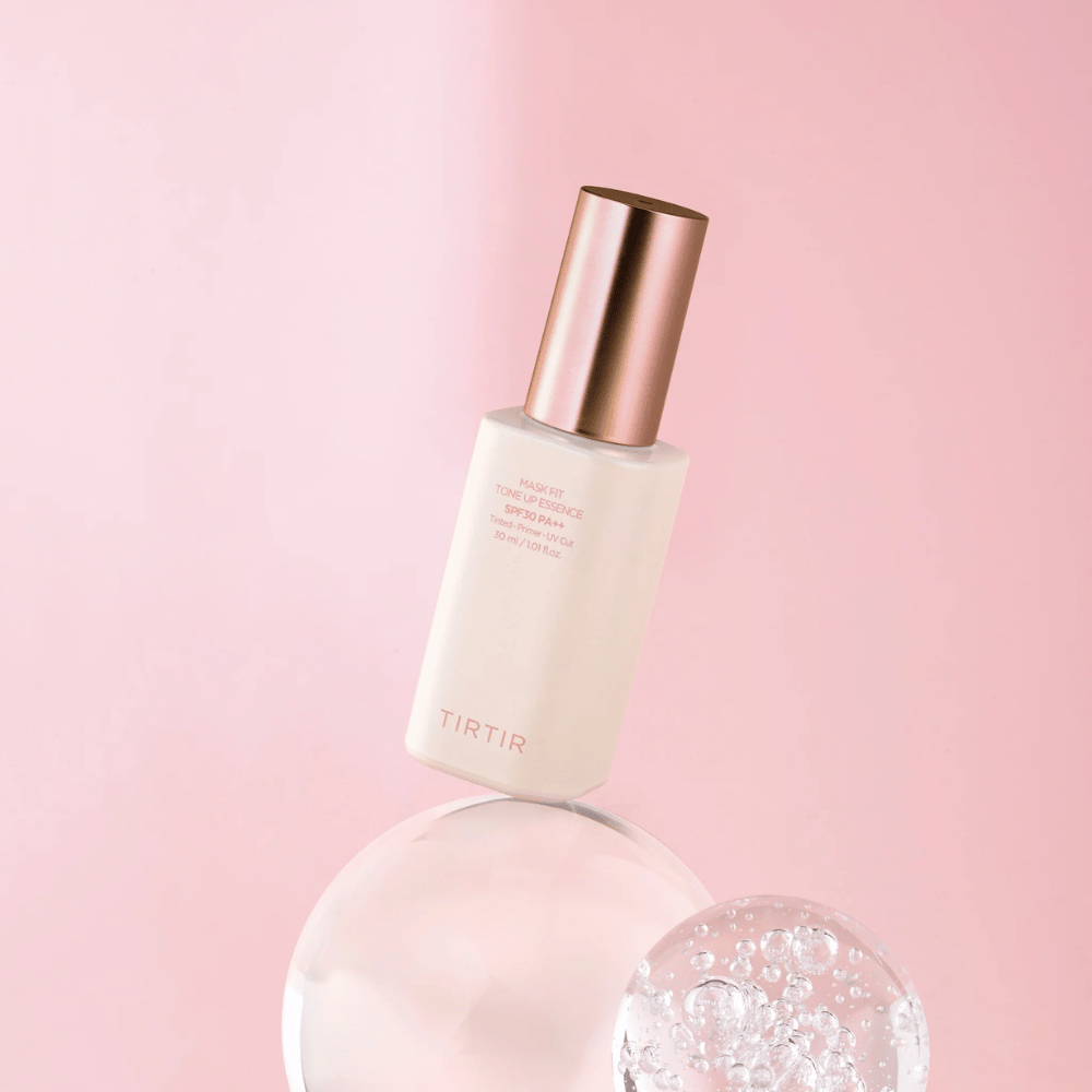 A imagem mostra uma garrafa de "TIRTIR MASK FIT TONE UP ESSENCE" com SPF30 PA++ contra um fundo rosa, que dá uma sensação de suavidade e feminilidade.