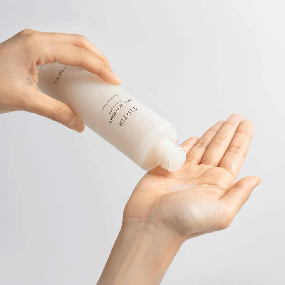 A imagem mostra uma pessoa derramando um líquido leitoso de uma garrafa marcada "TIRTIR Milk Skin" na palma da mão. O produto parece ser um tônico facial ou loção hidratante.