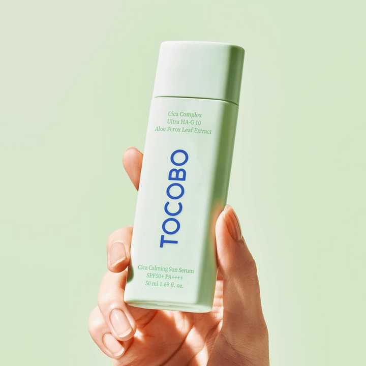 Hand håller i TOCOBO Cica Calming Sun Serum med mintgrön förpackning och blå text mot ljusgrön bakgrund.