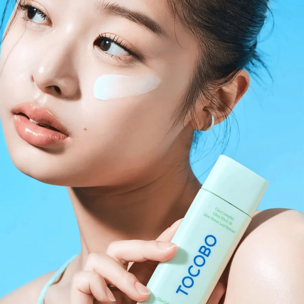 Modelo com protetor solar sob o olho segurando TOCOBO Cica Calming Sun Serum em embalagem verde menta contra fundo azul.