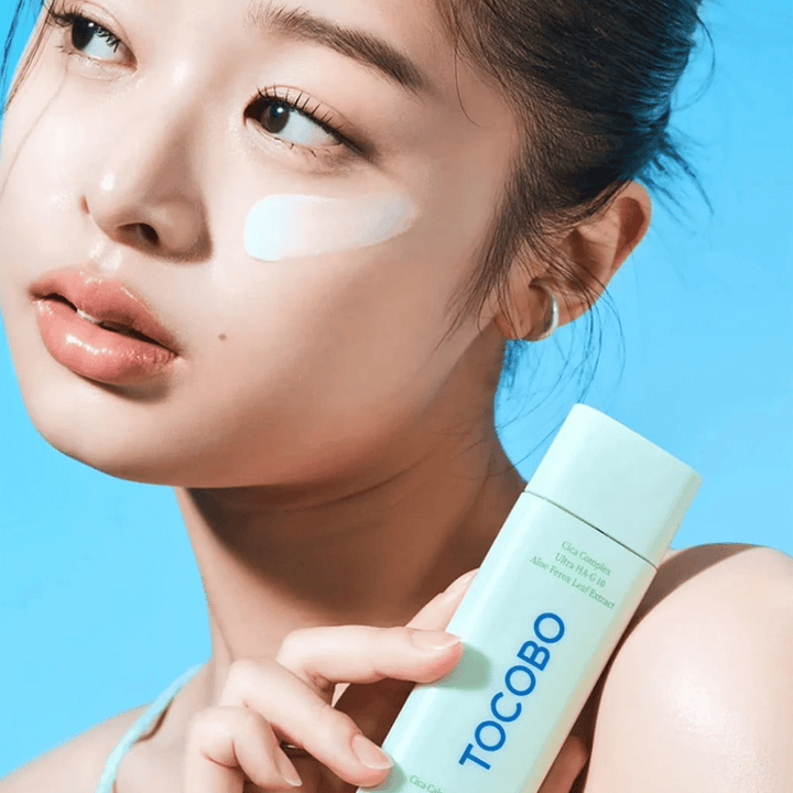 Modell med solkräm under ögat håller i TOCOBO Cica Calming Sun Serum i mintgrön förpackning mot blå bakgrund.
