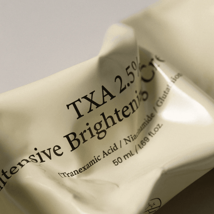 Close-up de AXIS - Y TXA 2.5% Intensive Brightening Cream, em que o design suave e minimalista da bisnaga está em destaque. A imagem realça o nome do produto, Tranexamic Acid e Niacinamide, bem como a elegante embalagem bege.