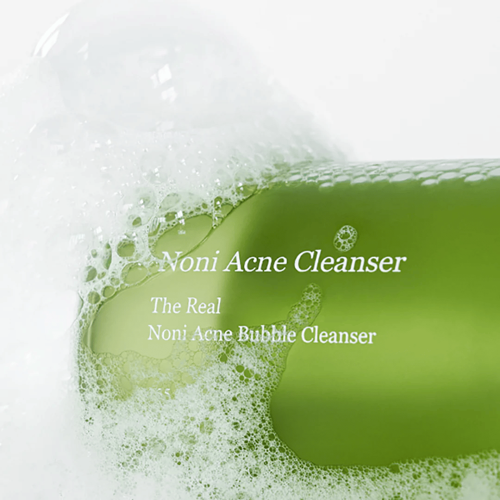 A garrafa com The Real Noni Acne Bubble Cleanser está em foco e rodeada por espuma abundante. A cor verde fresca sinaliza ingredientes naturais, perfeita para limpar e combater a acne. Um produto ideal para uma pele limpa e clara.