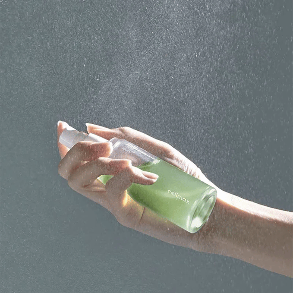 Uma mão segura Celimax Noni Mist frasco spray e aplica o produto em uma névoa fina e uniforme na pele. A garrafa é verde e o produto é comercializado como "The Real Noni Energy Ampule Mist" para fornecer energia e hidratação à pele.