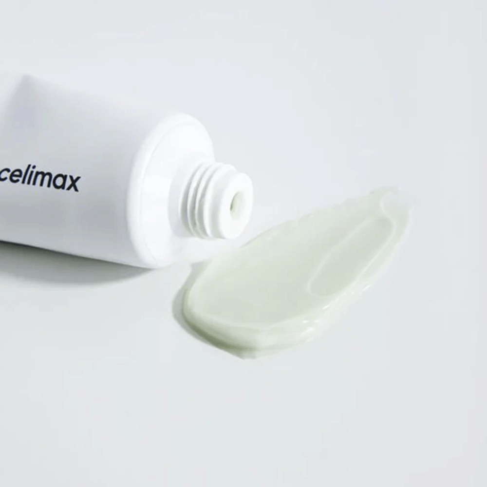 Celimax Noni Energy Repair Cream em um tubo branco com uma pequena quantidade de creme verde claro espalhado ao lado do tubo em uma superfície branca. O creme é para nutrir e reparar a pele.