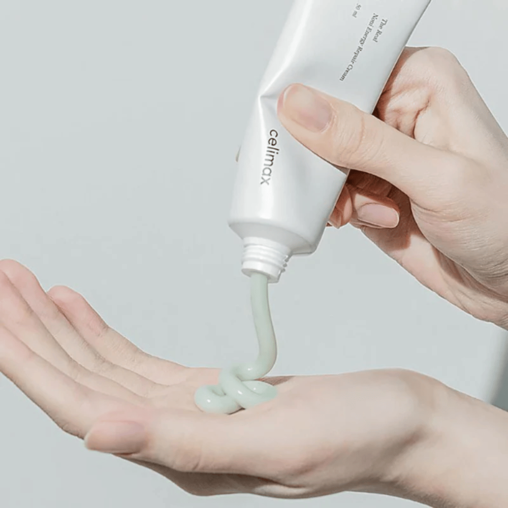Uma mão segura um tubo branco com Celimax Noni Energy Repair Cream, e o creme é pressionado em um jato uniforme na palma da mão. O creme é verde claro e é comercializado para nutrir e restaurar a pele.