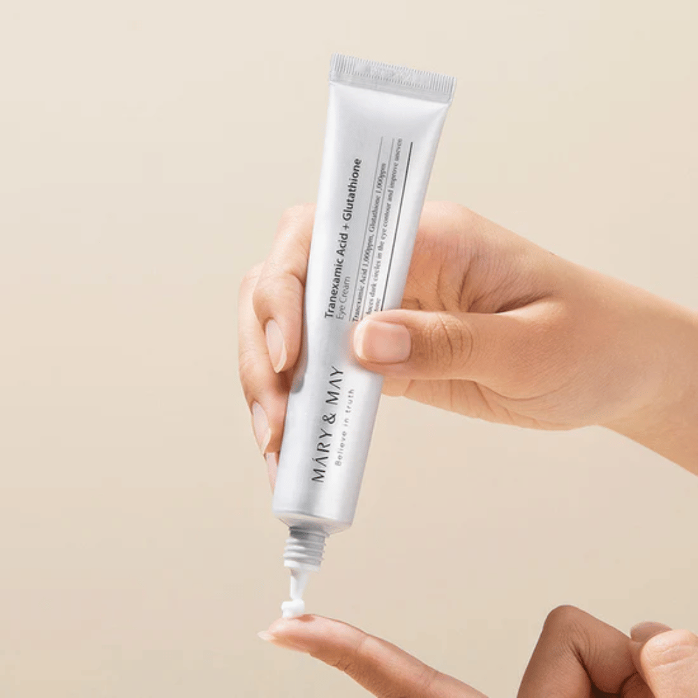 Uma mão segura um tubo MARY & MAY "Tranexamic Acid + Glutathione Eye Cream". O tubo é branco e o design é limpo e simples, destacando as propriedades calmantes e iluminadoras do produto para a área dos olhos. O produto é aplicado cuidadosamente, com foco na precisão e eficiência.