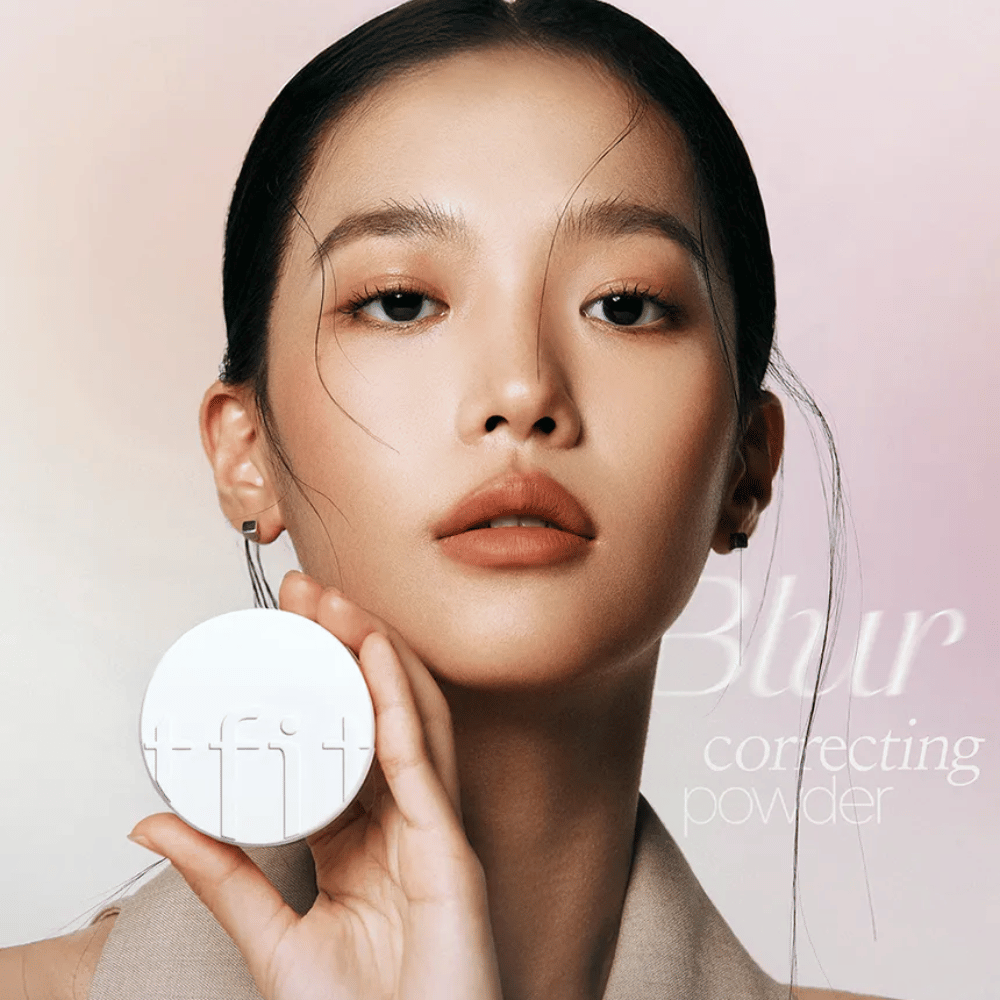 Mulher com maquiagem impecável segura uma caixa redonda branca com o logotipo TFIT, com o texto "Blur Correcting Powder" ao fundo.