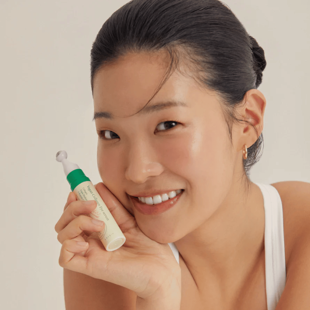 Uma mulher com cabelo preso e brincos dourados segura um AXIS-Y Vegan Collagen Eye Serum 10ml perto do rosto, enquanto sorri suavemente. O sérum é projetado para hidratar e reduzir rugas ao redor dos olhos, especialmente desenvolvido para pele sensível.