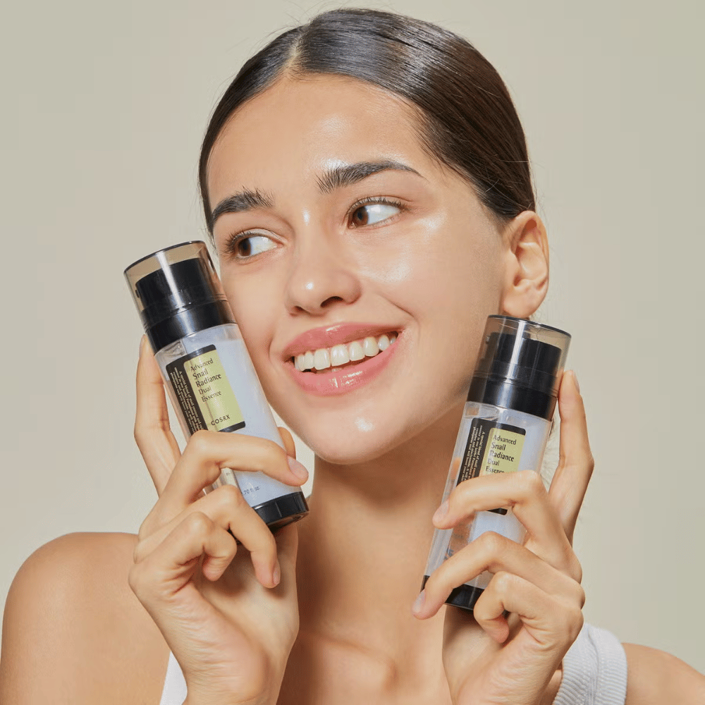 Mulher sorridente com pele brilhante segura duas garrafas COSRX Advanced Snail Radiance Dual Essence 80 ml perto do rosto contra um fundo claro.