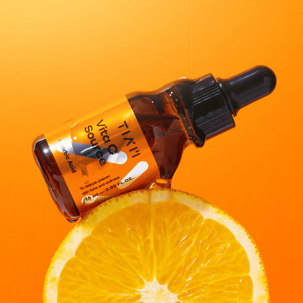TIA'M Vita C Source-frasco colocado contra uma laranja e um fundo laranja que simboliza a frescura e o efeito antioxidante da vitamina C.