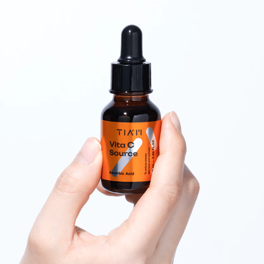 Mão segurando TIA'M Vita C Source, um sérum de vitamina C em frasco de vidro castanho com pipeta. O produto é projetado para uniformizar o tom da pele e dar brilho.