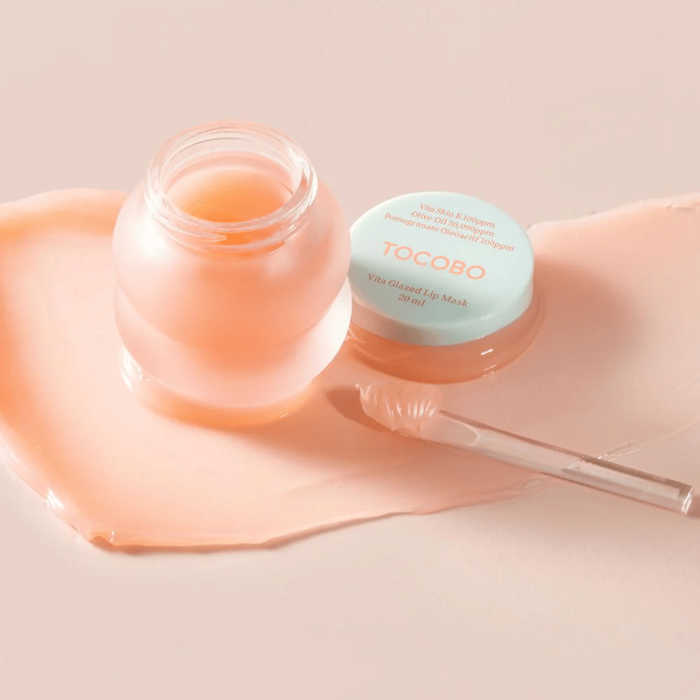 Um frasco com "Vita Glazed Lip Mask" de Tocobo é exibido. O frasco é transparente com uma tampa turquesa clara e contém uma máscara labial rosa e brilhante. Uma pequena espátula está ao lado do frasco, e parte da máscara está espalhada sobre a superfície. O produto é projetado para hidratar e cuidar dos lábios.