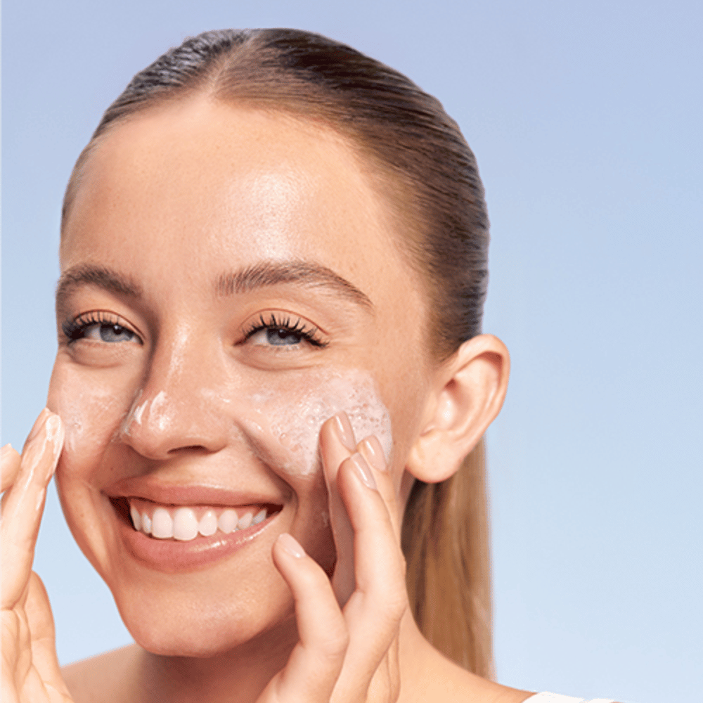 Mulher sorridente aplica LANEIGE Water Bank Gentle Gel Cleanser em forma espumosa nas bochechas, contra um fundo azul.
