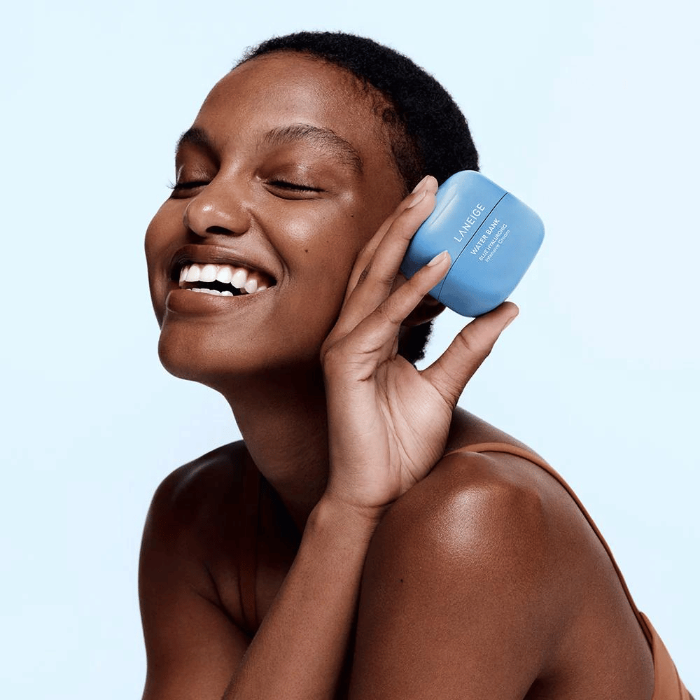 Mulher sorridente segurando uma lata azul de LANEIGE Water Bank Blue Hyaluronic Cream contra a bochecha, com fundo azul claro.