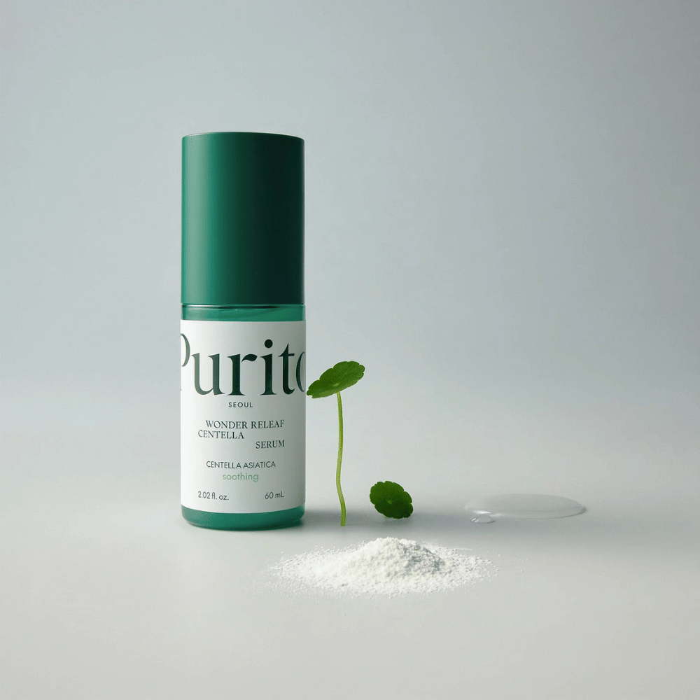 Garrafa verde de Purito Wonder Releaf Centella Serum 60 ml está sobre uma base branca, cercada por folhas de centella, um soro transparente em gotas e um pequeno pó branco.