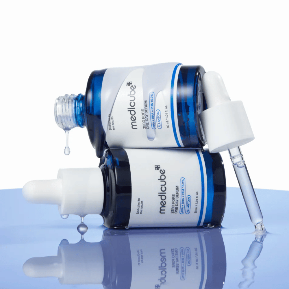 Duas garrafas azuis de Medicube Zero Pore One Day Serum deitadas uma contra a outra, com conta-gotas e gotas de soro sobre uma superfície azul refletora.