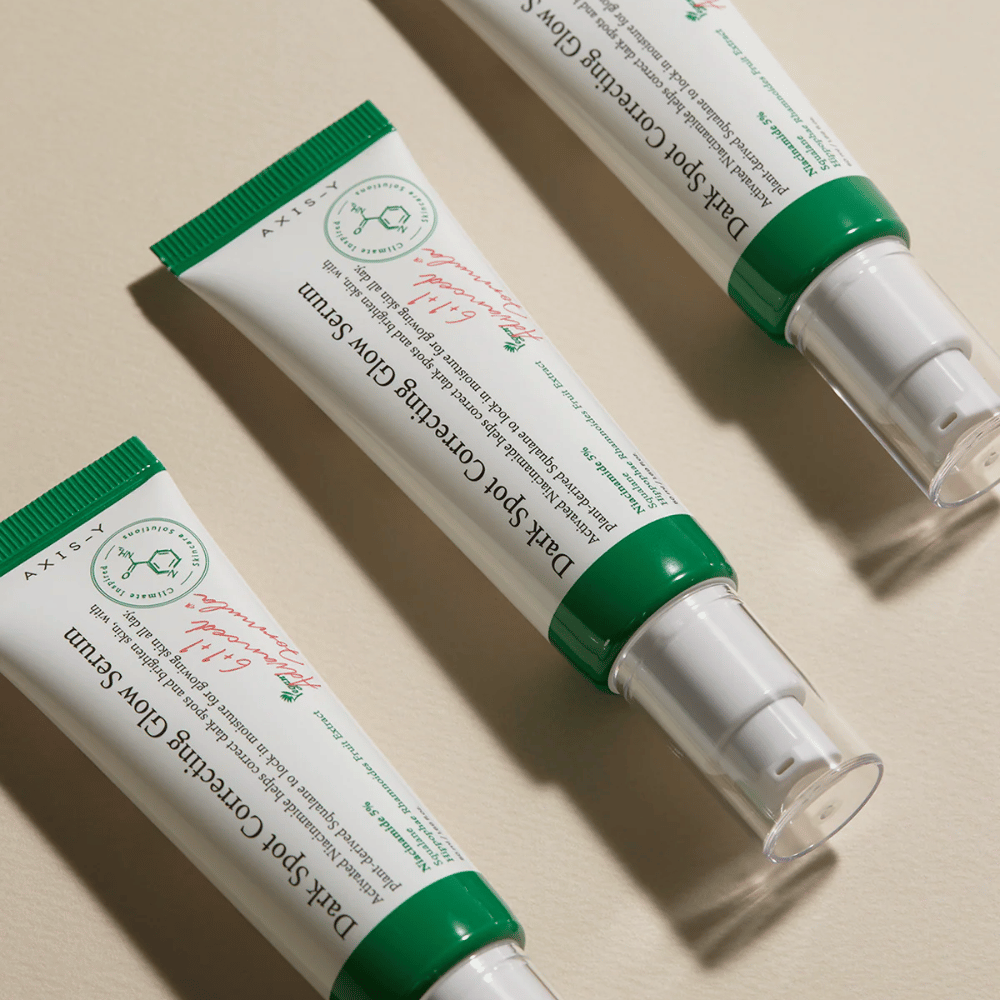 Três tubos de Axis-Y Dark Spot Correcting Glow Serum com tampa verde e embalagem branca estão sobre uma superfície clara.