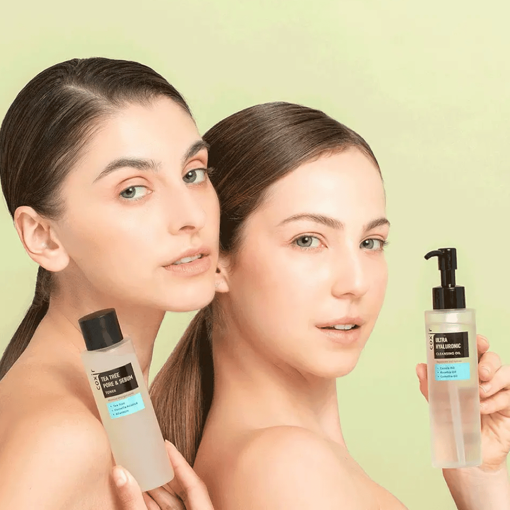 Duas mulheres com ombros nus posam com produtos de cuidados com a pele; uma segura um frasco rotulado "TEA TREE PORE & SEBUM", e a outra um frasco de bomba rotulado "ULTRA HYALURONIC CLEANSING OIL". Elas estão em frente a um fundo verde claro.