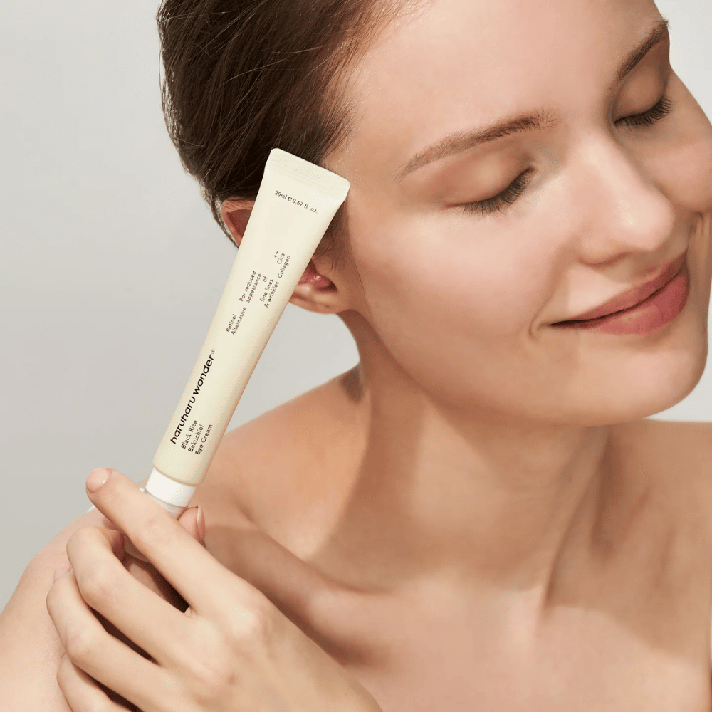 Uma pessoa sorridente com os olhos fechados segurando um tubo Haruharu Wonder Black Rice Bakuchiol Eye Cream contra a têmpora.