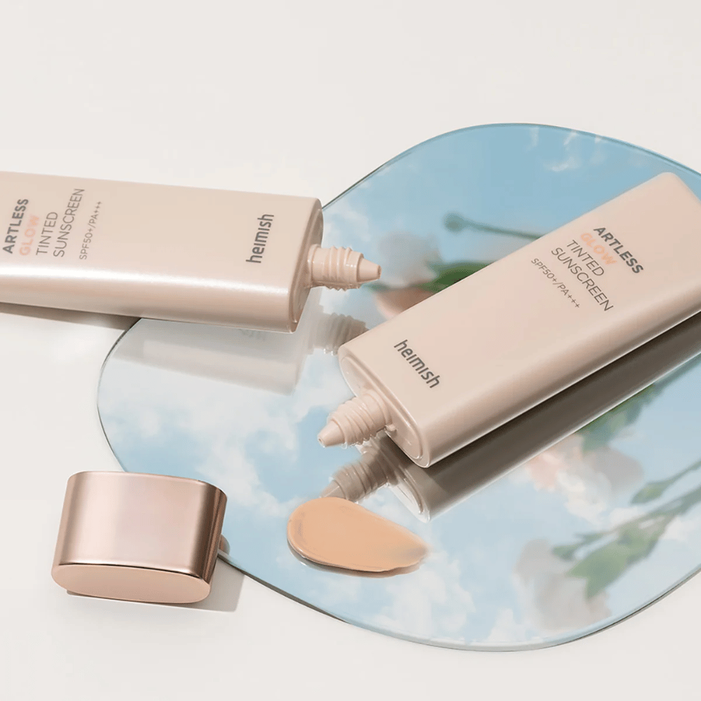 Uma garrafa heimish Artless Glow Tinted Sunscreen SPF50+ PA+++ 40ml e uma garrafa concealer em um espelho, que mostra diferentes tons de pele.