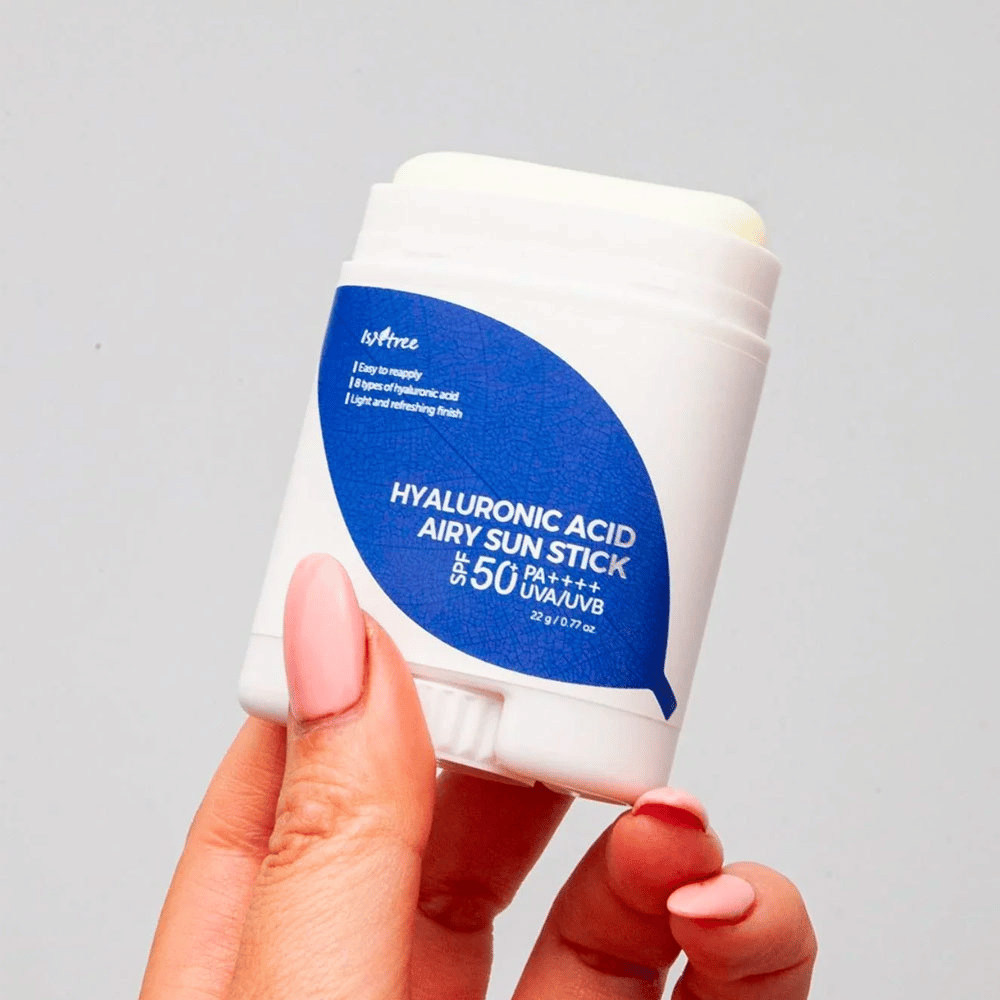 Uma mão segura um bastão branco de protetor solar com rótulo azul da marca Isntree. O produto chama-se "Hyaluronic Acid Airy Sun Stick" e tem fator de proteção solar SPF 50+ PA++++.
