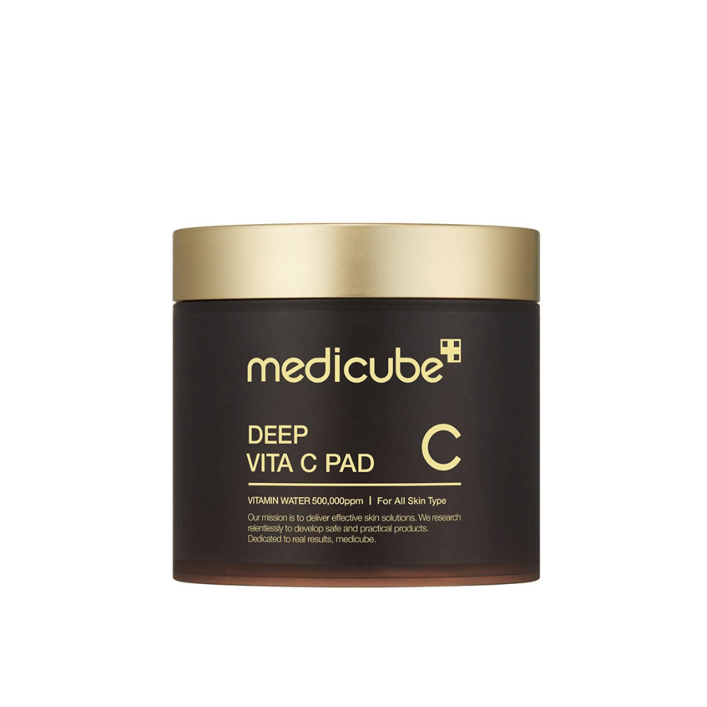 Um frasco de medicube, marcado "Deep Vita C Pad", com tampa dourada e corpo escuro. O texto no frasco indica vitamina C e adequação para todos os tipos de pele. O fundo é neutro.