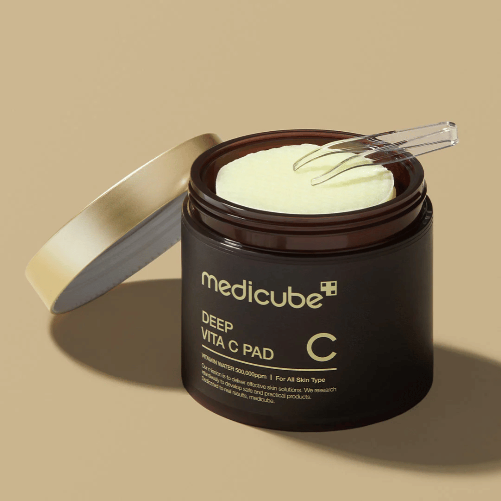 Um frasco com medicube Deep Vita C Pad. O frasco é marrom escuro com texto dourado e um rótulo branco invertido com informações sobre vitamina C. O interior da tampa é dourado, e uma pinça transparente está sobre os discos de algodão amarelo claro dentro. O fundo é de cor bege sólida.