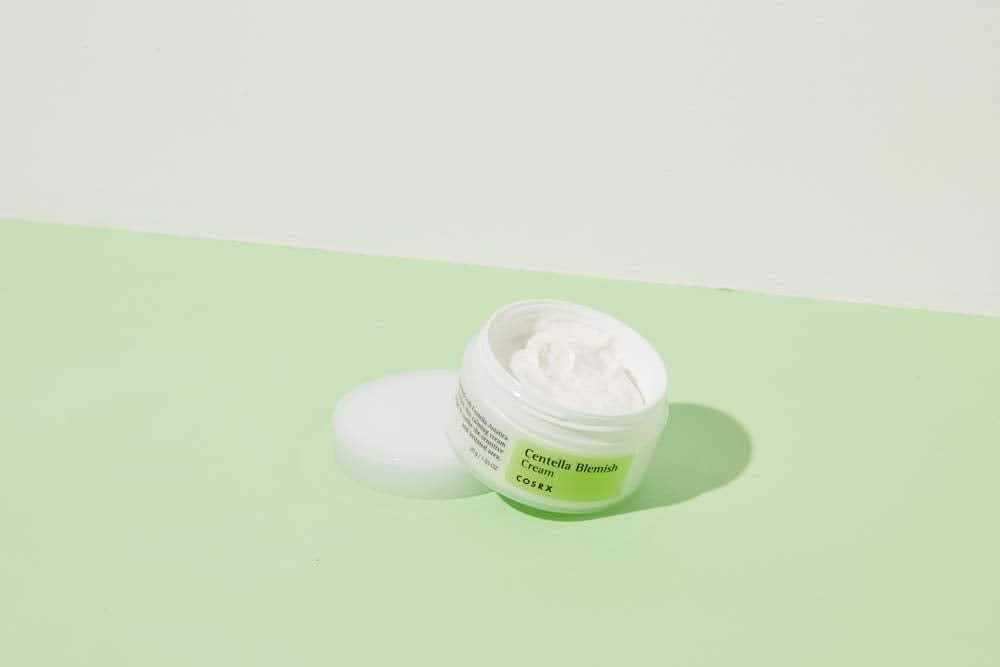 Um frasco COSRX Centella Blemish Cream 30ml que está sobre uma superfície verde.
