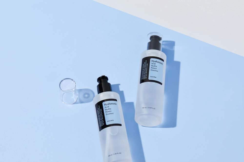 COSRX:s Hyaluronic Acid Hydra Power Essence 100ml proporciona benefícios hidratantes em superfície azul.