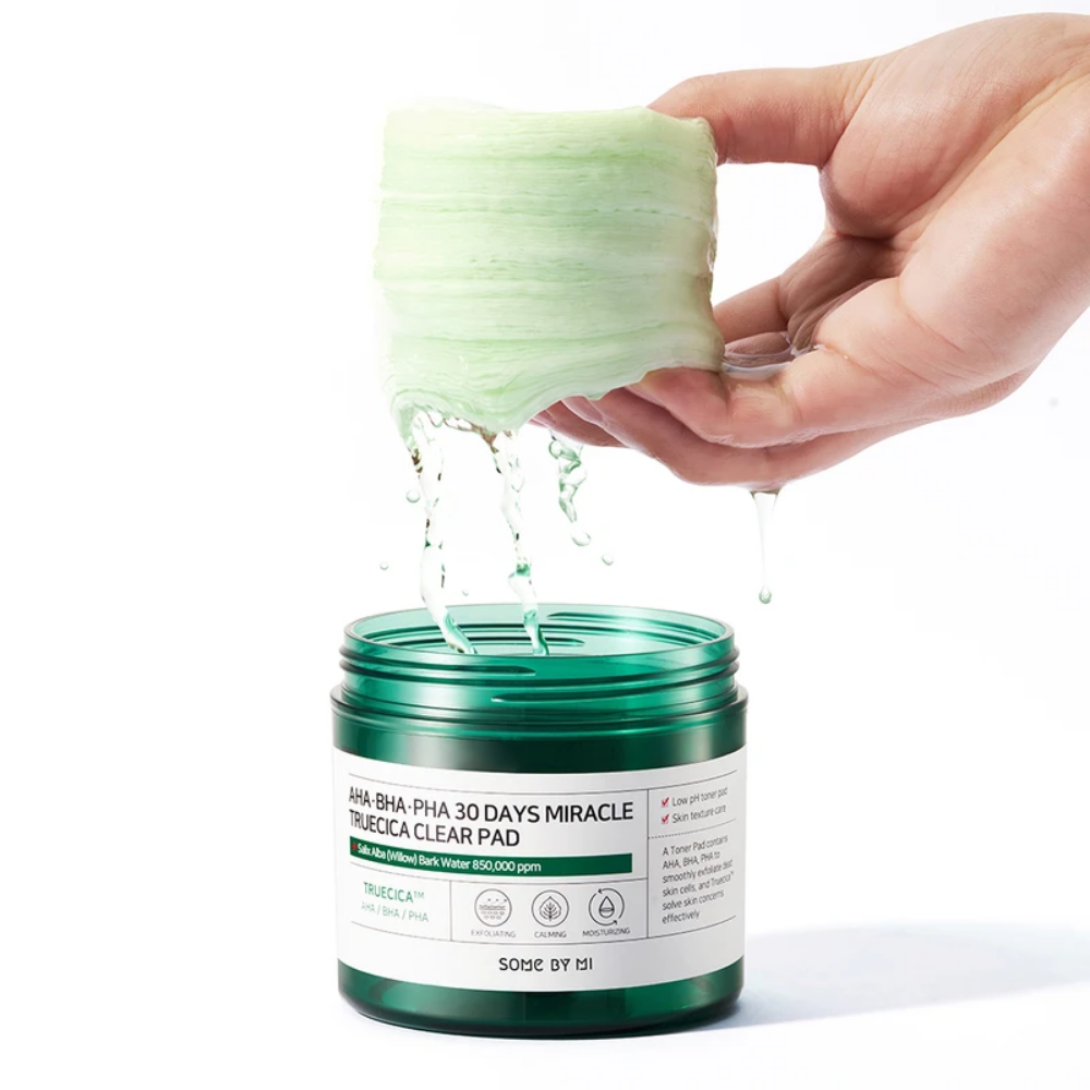 Uma mão segura um líquido verde em um frasco para SOME BY MI:s AHA, BHA, PHA 30 Days Miracle Truecica Clear Pad 70 unidades de tratamento de surtos de acne.