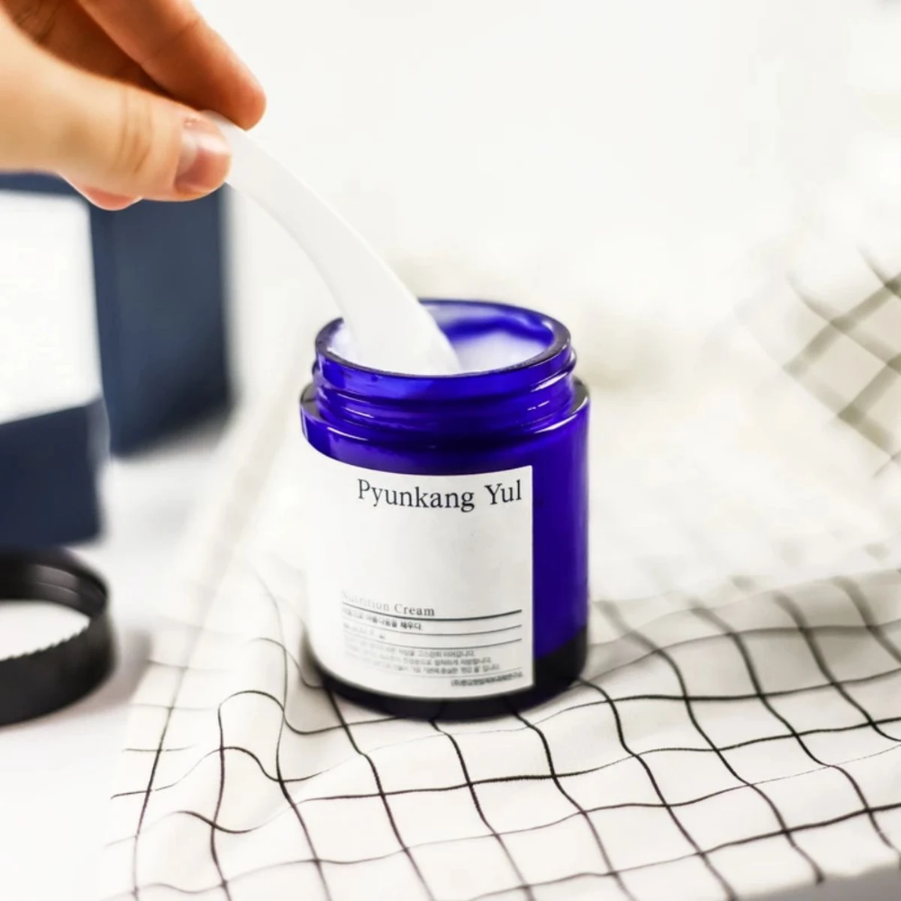 Uma pessoa usa um frasco Pyunkang Yul Nutrition Cream 100ml sobre uma mesa.