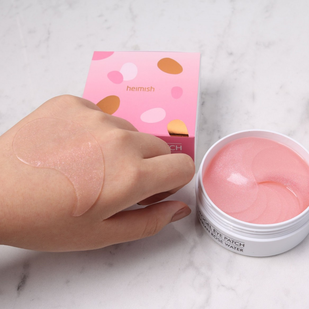 Uma mão segurando uma Bulgarian Rose Hydrogel Eye Patch rosa 60st de heimish e aplicando máscara para os olhos.
