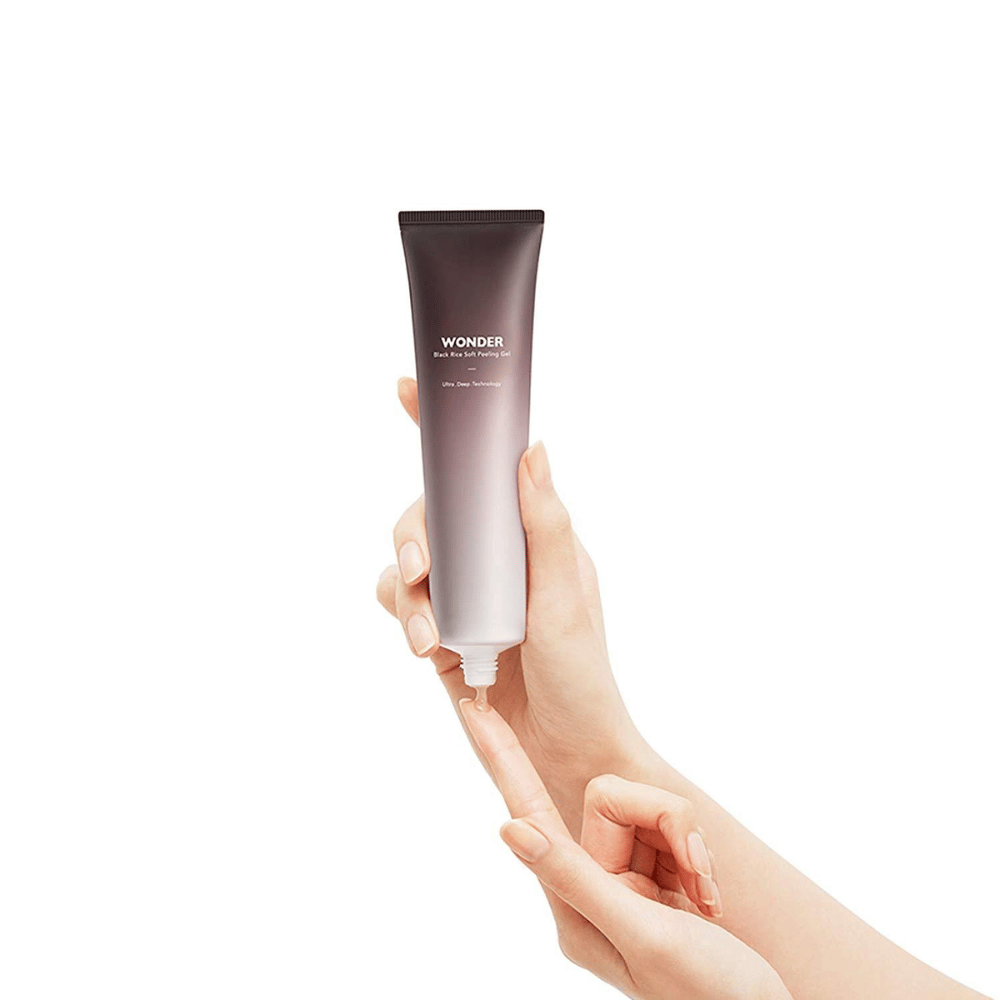 A mão de uma mulher segurando um tubo Haruhuru Wonders Black Rice Soft Peeling Gel 100 ml sobre um fundo branco.