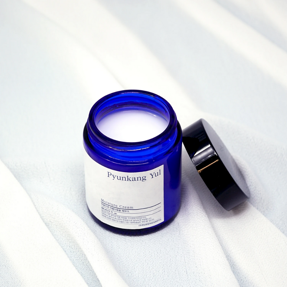 Um frasco azul com tampa em cima de um pano branco que exibe as propriedades hidratantes de 100ml de Pyunkang Yul's Moisture Cream.