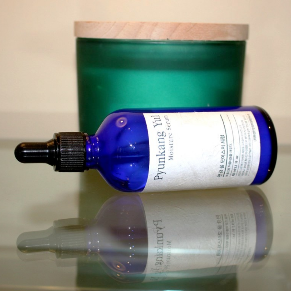 Uma garrafa Pyunkang Yul Moisture Serum 100ml equilibra os níveis de umidade em uma mesa ao lado de uma vela.