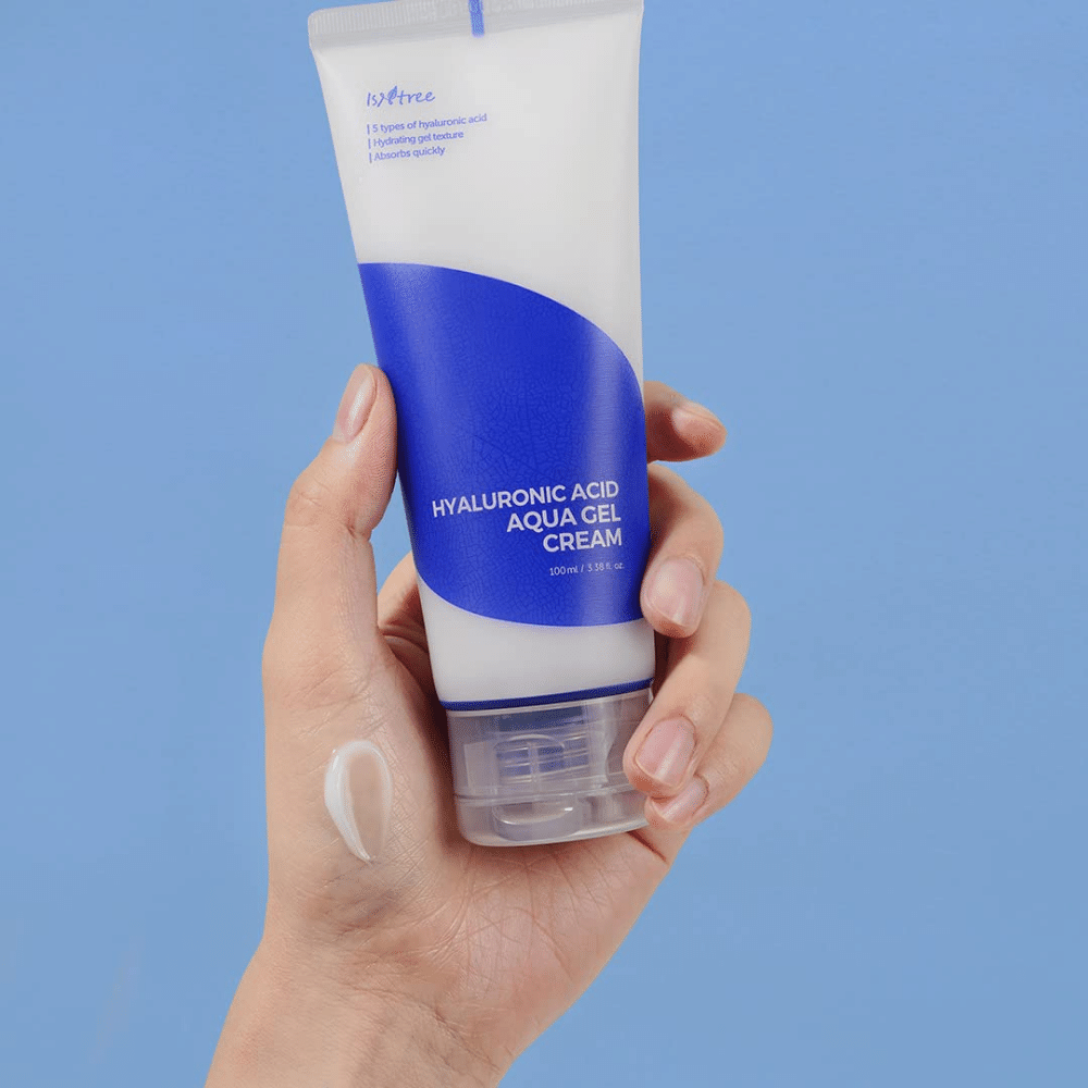 Uma mão segurando um tubo Isntree Hyaluronic Acid Aqua Gel Cream 100ml sobre um fundo azul.